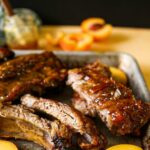 Maple peach bourbon ribs, ribs on a tray with sliced peaches, maple bourbon sauce behind it, bbq'd ribs on a platter