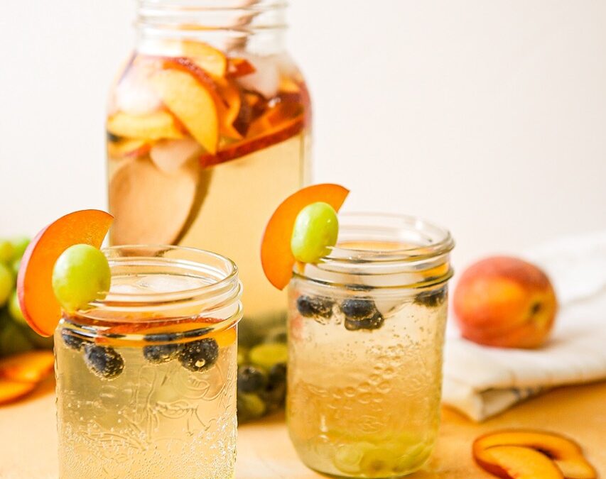 A large mason jar and two glasses of white wine and elderflower sangria. Fresh BC peaches and blueberries with green grapes.