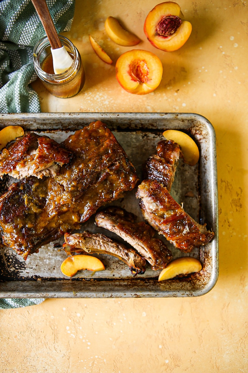 Ribs on a platter with fresh peaches, a sauce jar, and more peaches.