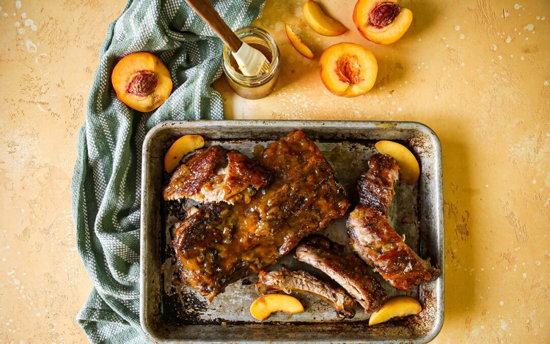 Baby backribs on a platter iwth fresh peach slices and a jar of sauce on the side with more peaches