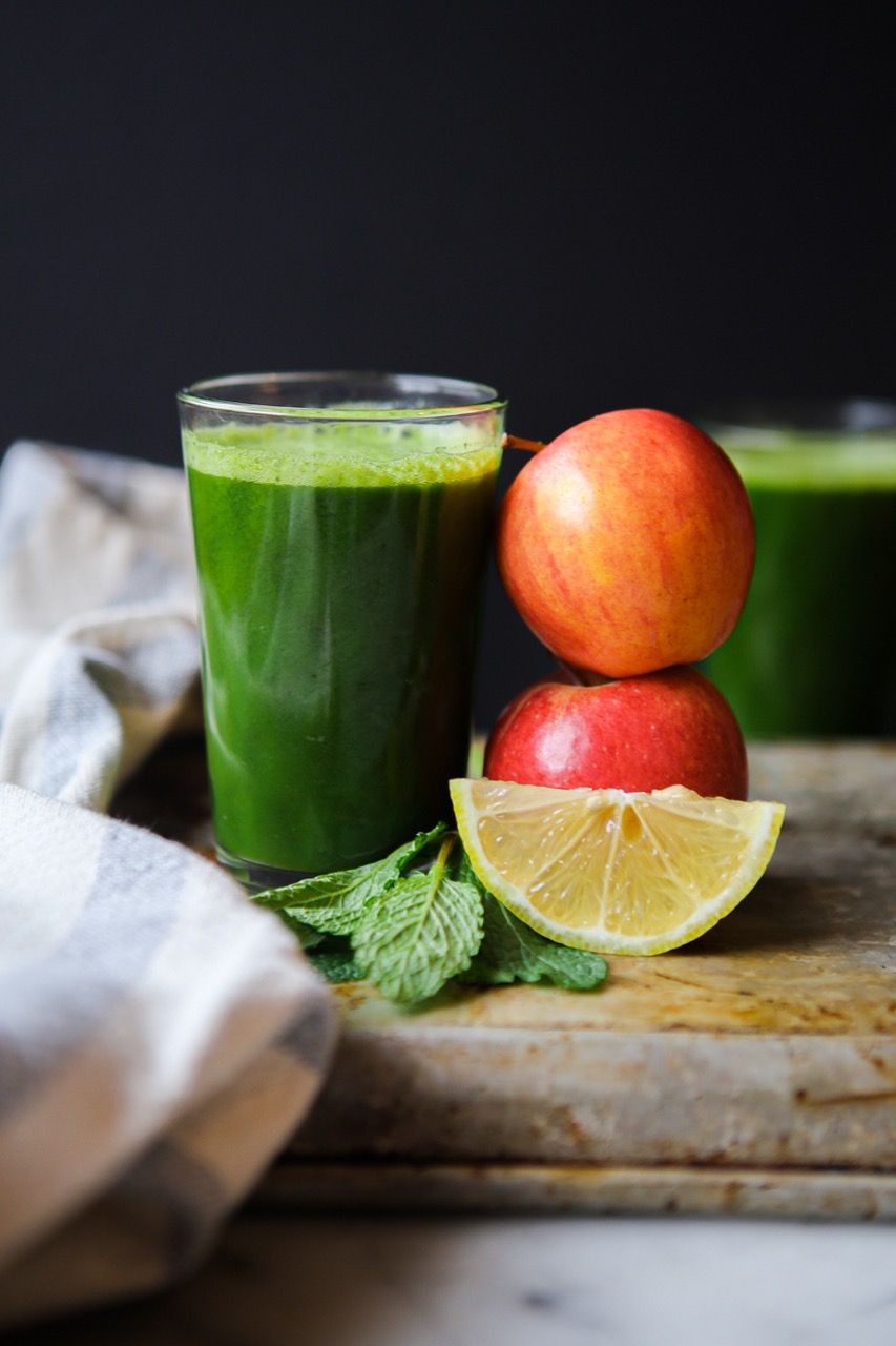 large glass of green juice wit apples stacked on top and a fresh lemon slice with mint leaves.