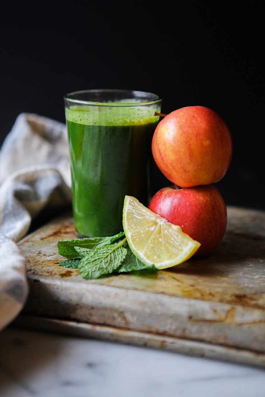 Green juice in a large glass with fresh crisp apples, lemon slices and mint leaves next to it.