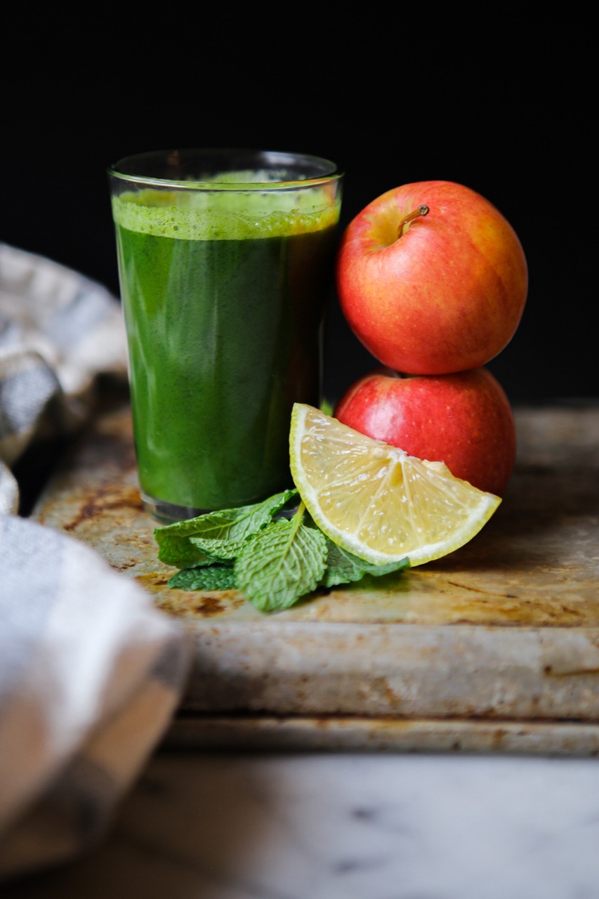 a large glass of green juice with fresh mint and lemon slices. Two apples stacked one on top of the other next to it.