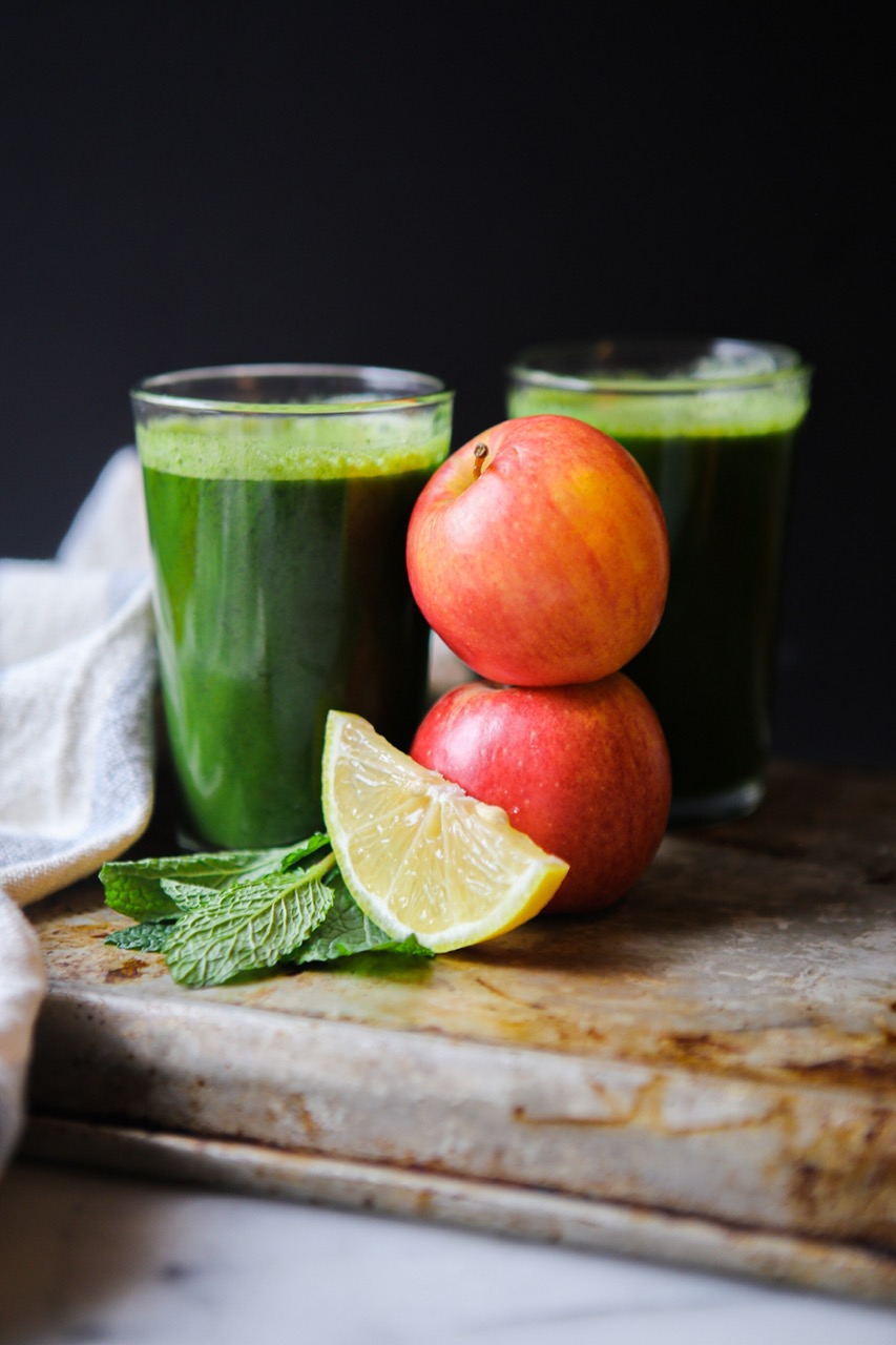 two glasses of vibrant green juice, one in front of the other with fresh apples, lemon slices and mint.