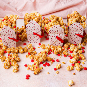 four little white takeout boxes with red and pink hearts filled with cinnamon heart and caramel popcorn